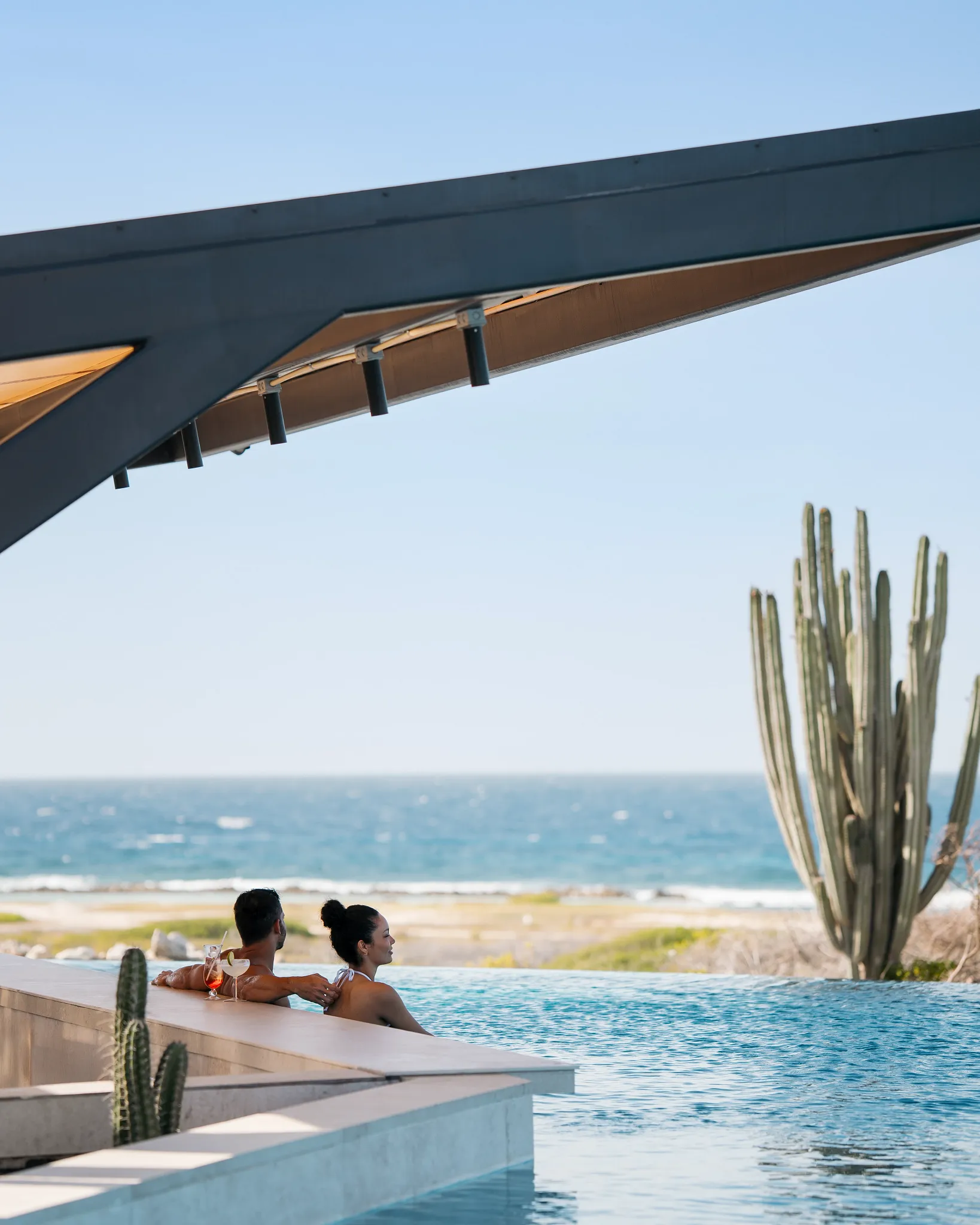 Couple relaxing at an infinity-edge pool with tropical drinks overlooking Baby Beach in Aruba.