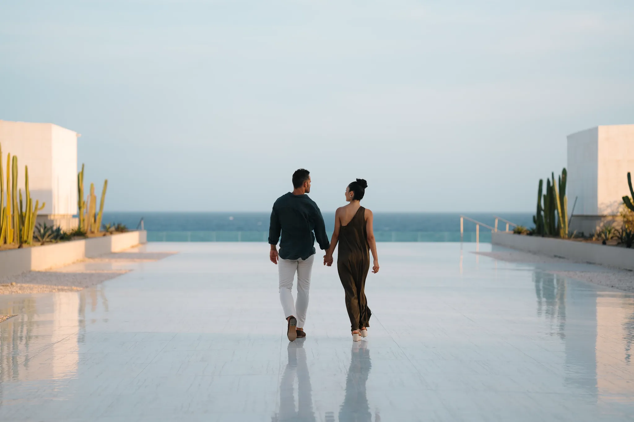Couple holding hands walking across a modern oceanfront terrace at Secrets Baby Beach Aruba.