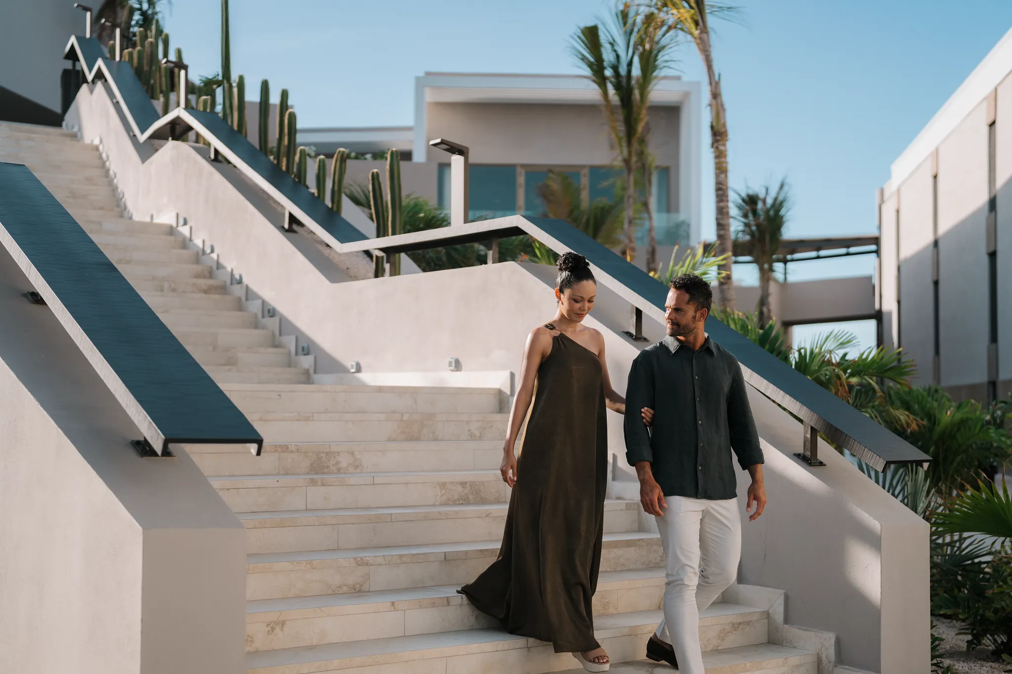 Stylish couple walking down modern resort steps at Secrets Baby Beach Aruba surrounded by tropical landscaping.